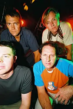 Three members of the band are seen. Two guitarists are facing away from the audience: towards the drummer who is between them at the back of the stage. The one at left wears a red, checked shirt over dark blue jeans. The drummer wears a dark cap and dark t-shirt; he is facing forward and slightly to his left. The second guitarist wears a light shirt with a montage of images over light blue jeans. Beyond the musicians is stage equipment and speakers.