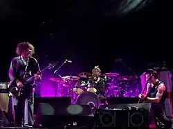 On a purple-lit stage, two men play guitars while a third man sits behind a drum kit.