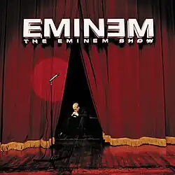 Image of a stage with a microphone stand and a spotlight pointing to where a performer would be. There is no performer seen. Curtains are slightly opened to reveal a man seated on a low chair. He is wearing a suit and his head is facing down. His hands are on his mouth to indicate worry. At the top, sits a 3D version of the Eminem logotype. Below it is text that reads "The eminem show" in a smaller font