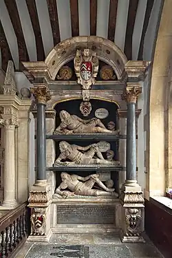 This image shows a grand multi-tiered Renaissance monument featuring three recumbent male figures in Roman armour lying on separate stone shelves within an elaborate architectural framework. The monument is constructed with classical elements including Corinthian columns, ornate capitals, and a decorative arched canopy topped with heraldic shields. The three effigies are positioned one above the other, each reclining on individual stone slabs with their heads resting on pillows. The architectural surround is made of light-coloured stone with darker accents, and the overall composition rises impressively within what appears to be a church interior with exposed wooden ceiling beams visible above. At the base is a large inscribed memorial tablet, and heraldic shields appear at various points throughout the structure, indicating this commemorates members of an important noble family.