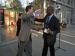 A white man, Gavin Newsom, wearing a gray suit reaches to embrace Jones, while holding a book in his right hand. Jones, who is also reaching out, wears a dark suit and has a microphone and piece of paper in his left hand. Inside a glass-walled building behind them, a display says "Climate is an angry beast and we are poking at it with sticks".