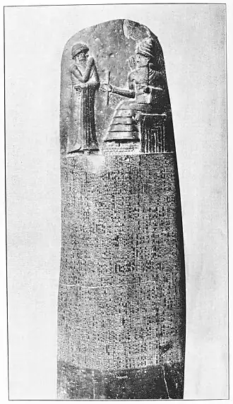 A stone monument with two parts; at top, a relief depicting two figures, one standing and one seated; at bottom, cuneiform text of the Hammurabic legal code of ancient Babylon.