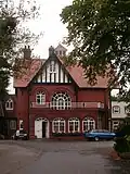 A red brick house, at the end of a long drive, seen from the south. Prominent is a gable with black and white striped pebbledash.