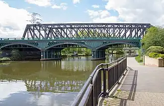 A colour photo of grey and blue three-arch viaduct slightly obscured on the right by a tree. It is crossing a river and there is a steel truss bridge immediately behind it.