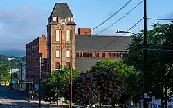 A brick tower with peaked roof on the right-hand side of a street in front of the camera. There are green hills in the background.