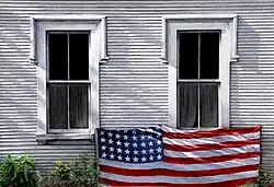 oil painting of a white clapboard house, two windows with flag draped along bottom sills, weeds along base