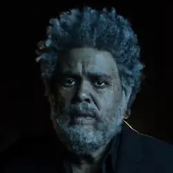 Aged and bearded the Weeknd in a dark setting with a small ray of light shining over his shoulder