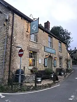 The image shows The White Lion pub in Broadwindsor at sunset. The front entrance is shown with the green sign in the centre of the image