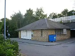 A single-storey square yellow-brick building viewed from a 45 degree angle with windows on either side. The photo is taken from across the road with the platforms visible elevated behind the building.