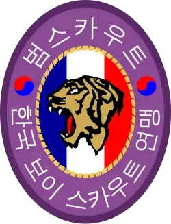An oval badge of red with 범스카우트 (Tiger Scout) and 한국 보이 스카우트 연맹 inscribed in hangul; The center is an oval of yellow with the profile of a tiger head over a scroll inscribed with "preparation" and knot