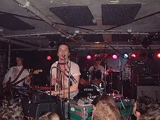 A band play in a crowded room. A man is playing keyboards while singing into a microphone; behind him is a drummer and to one side is a guitar player.