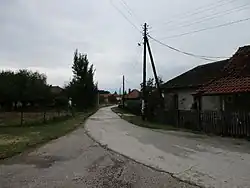 The image shows a part of Trbunje. You see a concrete road. On the left side of the road, you see a barbed-fence behind which stand three trees. You can see the top of a red roof in the distance behind the trees. On the right side of the road, there is an electricity pole and a wooden fence. Behind the wooden fence, you see two houses right next to each other. One with gray outer-walls and black slanting roof. The roof must have been red at some point. There are two windows on the outer-wall. The second house has a slanting red roof that goes past the doorway, creating a makeshift porch-area. You can see more houses and trees towards the end of the road. This whole scenery is set against the backdrop of a cloudy sky.