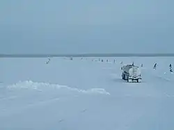 Large trucks, widely spaced, travel single-file across a frozen river.