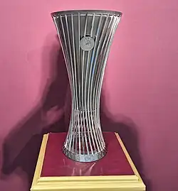 UEFA Europa Conference League Trophy. The trophy is standing on a claret coloured pedestal with gold trim, in front of a claret coloured backdrop.