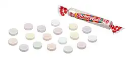 An image of Smarties tablet candies on a white background. On the right is a roll of candies consisting of 15 multicolored tablets in a clear film wrapper. The wrapper displays the Smarties Candy Company logo. On the left are a selection of loose Smarties candies.