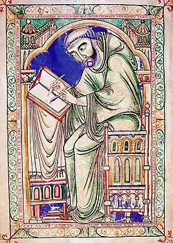 A man with a tonsure haircut in medieval robes sits at a table writing into a book with a quill.