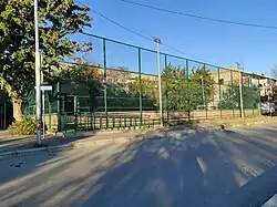 Corner view of an outdoor football (soccer) pitch, named after Vakhtang Pangani, on a sunny day. The pitch is enclosed by a tall, green chain-link fence and has green artificial turf. It is on a street corner, with long shadows cast across the pavement in the foreground. A blue plaque with the name is visible on a pole to the left, and residential buildings and trees are seen in the background under a clear blue sky.