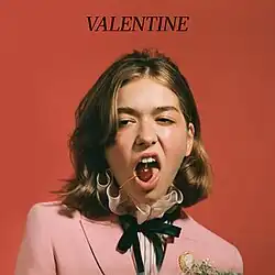 Cover image of "Valentine": a woman wearing a pink suit poses with a cherry in her mouth, in front of a dark red background. The name of the song in all caps, "VALENTINE", is printed above her.