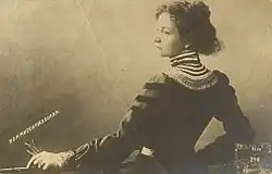 Monochrome photograph of a young woman in a high-necked 19th century dress. Subject is shown from the waist up standing at a balcony and facing to the left