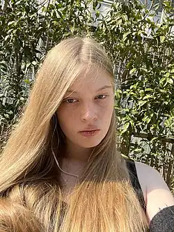 Selfie of Vivian Jenna Wilson. She is a young white woman with long blond hair wearing a black top, standing in a garden in view of the sun.