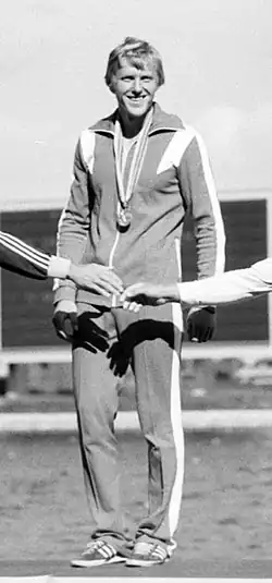 Vladimir Parfenovich standing in a track suit with a gold medal around his neck, while two hands reach out and touch each other in front of him.