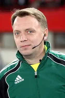 Portrait of a man wearing a dark green Adidas jacket over a neon yellow shirt. He has an earpiece on his left ear.