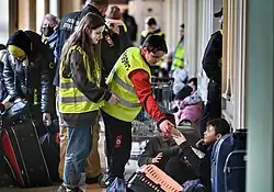 Two young people in hi-viz jackets hand out bottled drinks as other people pass by with luggage