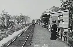 A lady standing on a station platform looks at the camera while a train pulls in from in the distance
