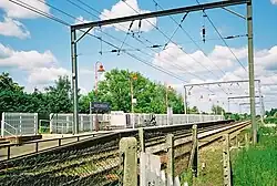 A view of a railway platform on the left-hand side of two tracks as seen from a parallel path slightly lower. On the platform there are red lampposts and a sign that reads "WATERBEACH". There are many overhead gantry structures and wire fencing obstructing the view of the tracks on the left-hand side.