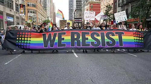 A crowd of 2019 protestors carry a variety of signs as they march behind a large rainbow-striped banner reading "We Resist"
