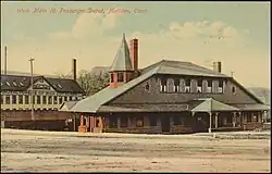 Postcard showing a two-story railroad station with a smaller second story. The roof from the second story continues down to the ends of the first story. A small turret is located on one end.
