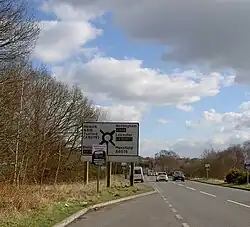 Large rectangular elevated road sign with white background and black markings depicting five spokes around a central round hub on a sunny day against a blue and white sky