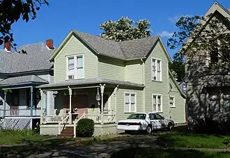 Photograph of a modest two-story wooden house with porch, closely flanked by other houses