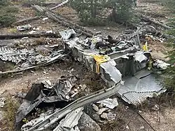 A piece of metal wreckage with bolts and rusted fasteners is positioned above a memorial plaque. The plaque reads, “In Loving Memory, Malory Kimmel, 1949–1970.” Behind the plaque, more crumpled metal wreckage is visible, with plants and rocks surrounding the area.