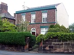 A sandstone wall with a hedge and a gate in front of a plain brick house in two storeys with three bays and an arched central doorway; the right side of the house is painted white and the edge of a larger house is to the left