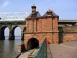 A red sandstone building seen from the rear, in two storeys, with an arch over a footpath on the left in the lower storey, and a plain wall in the upper storey; two chimneys and two hipped roofs; on the left are parts of the bridges and on right is the edge of the approach to the road bridge