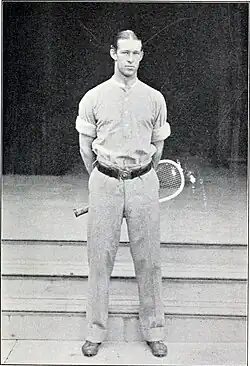 A man in white pants and shirt swings a wooden racket on a grass court, with other male and female tennis players in the background