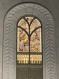 A stained glass window with branches depicting cherry blossoms and leaves, surrounded by gold. The window is surrounded by carved masonry, depicting branches and cherry blossoms stretching from the top of the window to the bottom along an arch.