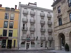 Photograph of a 5-storeys building with 4 windows per story, shot from a square. It has a 4-storeys building on the left. A street leaving the square separates the hotel from a representative building. The entrance of the hotel is crowned by a sign "HOTEL (four stars) ASTURIAS" and three flags