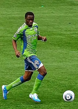 Steve Zakuani, dressed in a green jersey and blue shorts, runs towards a ball while playing soccer