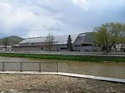 The inclosed ice stadium shown from the side with the river in the front on a cloudy but sunny day.