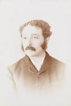 A man in early middle age, with a buttoned-up coat, sideburns and a bushy moustache.