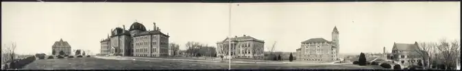Shows a black and white panorama of buildings on a campus in an open plain.